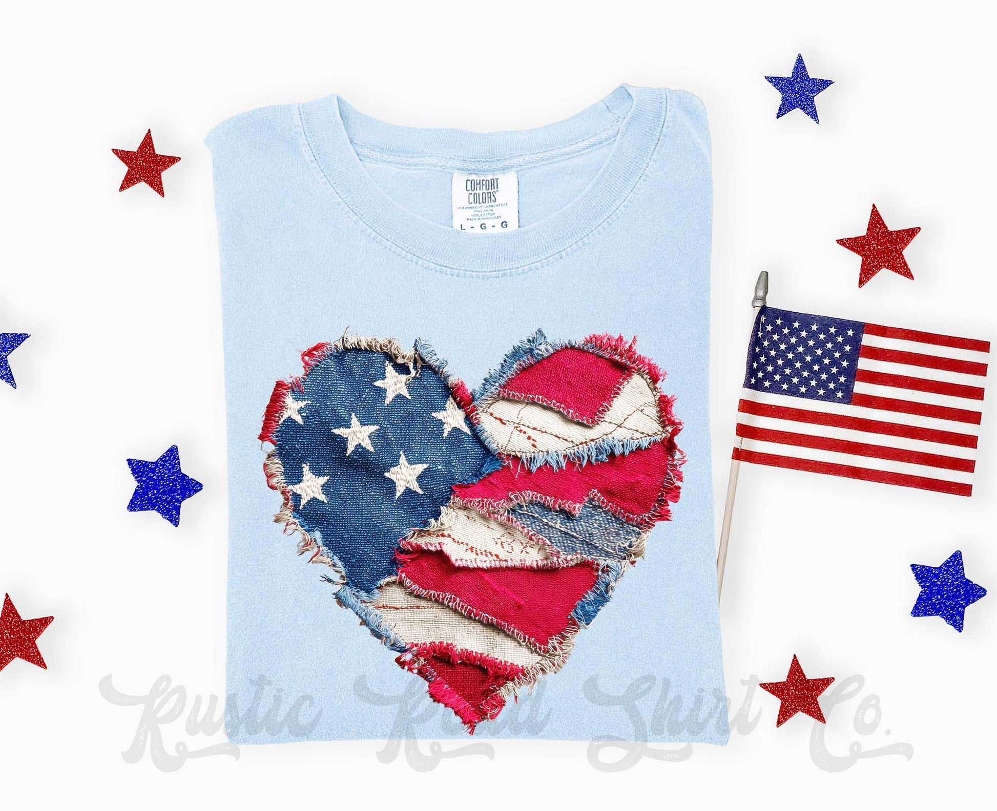 Light blue t-shirt with a heart-shaped American flag design, surrounded by small American flags and stars on a white background.