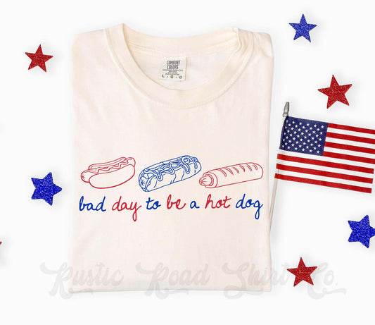 Folded white t-shirt with hot dog, bun, and sausage graphic and text 'bad day to be a hot dog' on a white background with stars and an American flag.
