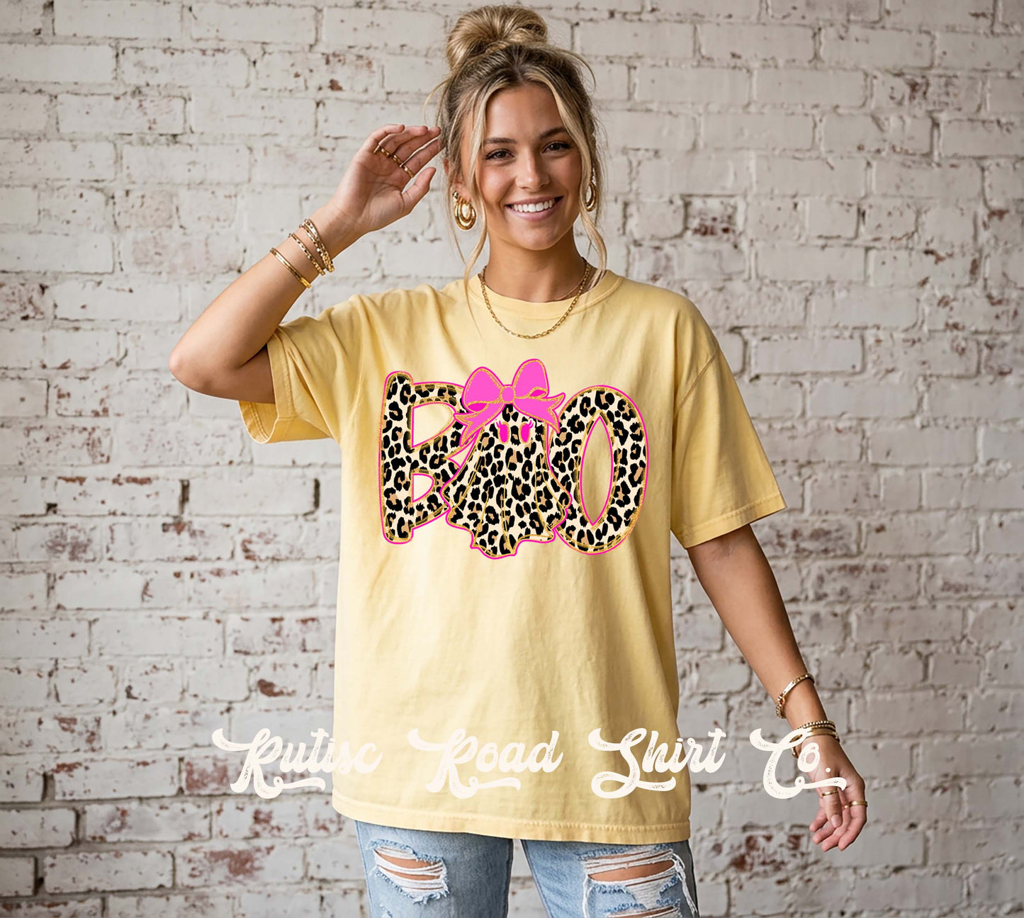 A woman wearing a pale yellow graphic t-shirt with a pink bow and leopard print 'BOO' design, standing against a white brick wall. A watermark for 'Rustico Road Shirt Co' is visible at the bottom.
