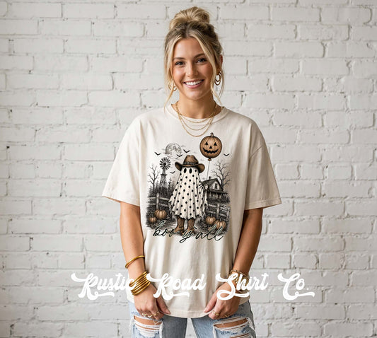 A woman wearing a cream-colored graphic t-shirt with a Halloween-themed illustration, standing against a white brick wall. The t-shirt features a spooky ghost, pumpkins, and the text 'Blessed & Beloved'.