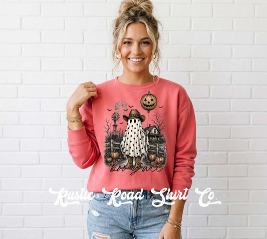 A woman wearing a coral pink crewneck sweatshirt with a detailed Halloween graphic print, paired with light-wash distressed jeans, standing against a white brick wall. A watermark for 'Krisie Road Shirts Co' is visible at the bottom.