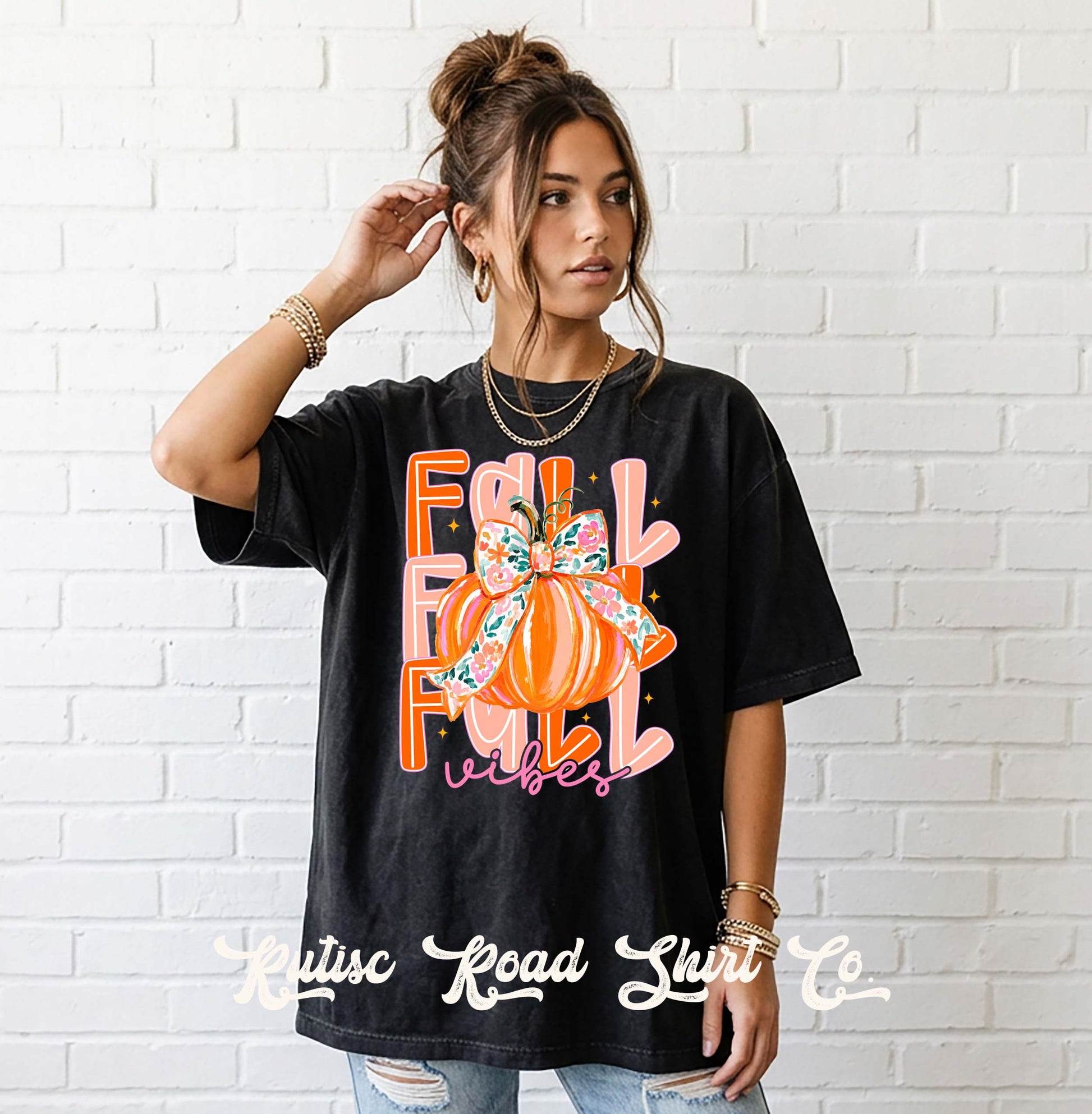 A woman wearing a black oversized graphic t-shirt with a colorful autumn-themed design that reads 'FALL FOLLY vibes' with a pumpkin and bow motif, posed against a white brick wall. The brand name 'Kitsuc Road Shirts Co.
