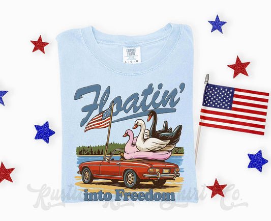 Light blue t-shirt with a graphic of a convertible car, American flags, and swans with stars around on a white background.