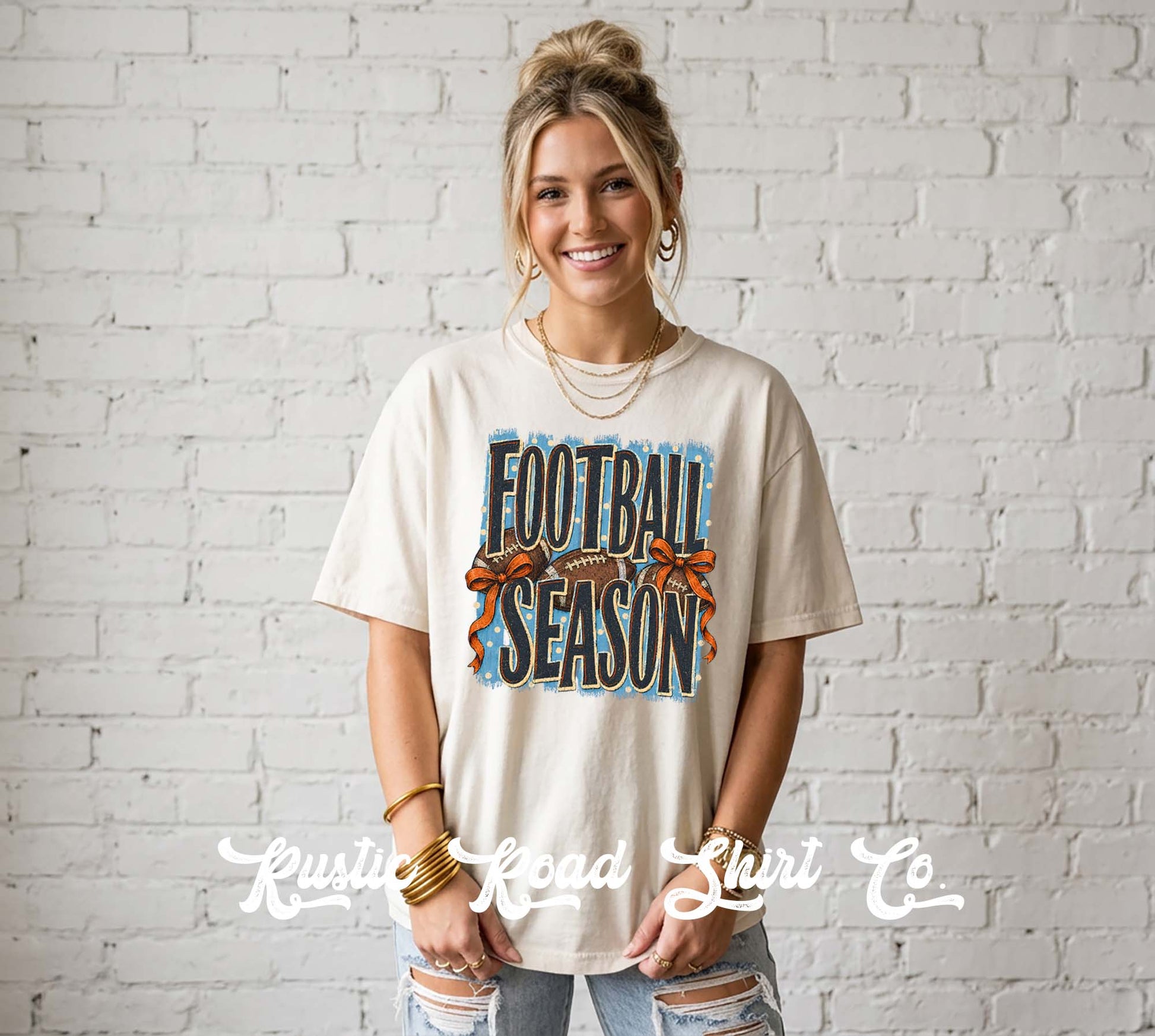 A woman wearing a cream-colored graphic t-shirt with 'FOOTBALL SEASON' printed in blue and orange, standing against a white brick wall. A watermark for 'Krusty Road Shirt Co' is visible at the bottom.