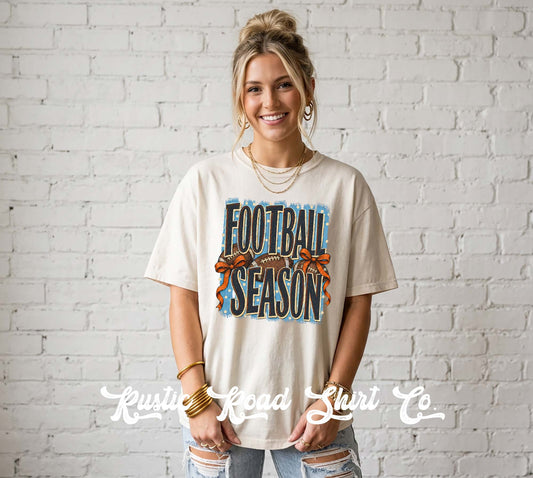 A woman wearing a cream-colored graphic t-shirt with 'FOOTBALL SEASON' printed in blue and orange, standing against a white brick wall. A watermark for 'Krusty Road Shirt Co' is visible at the bottom.