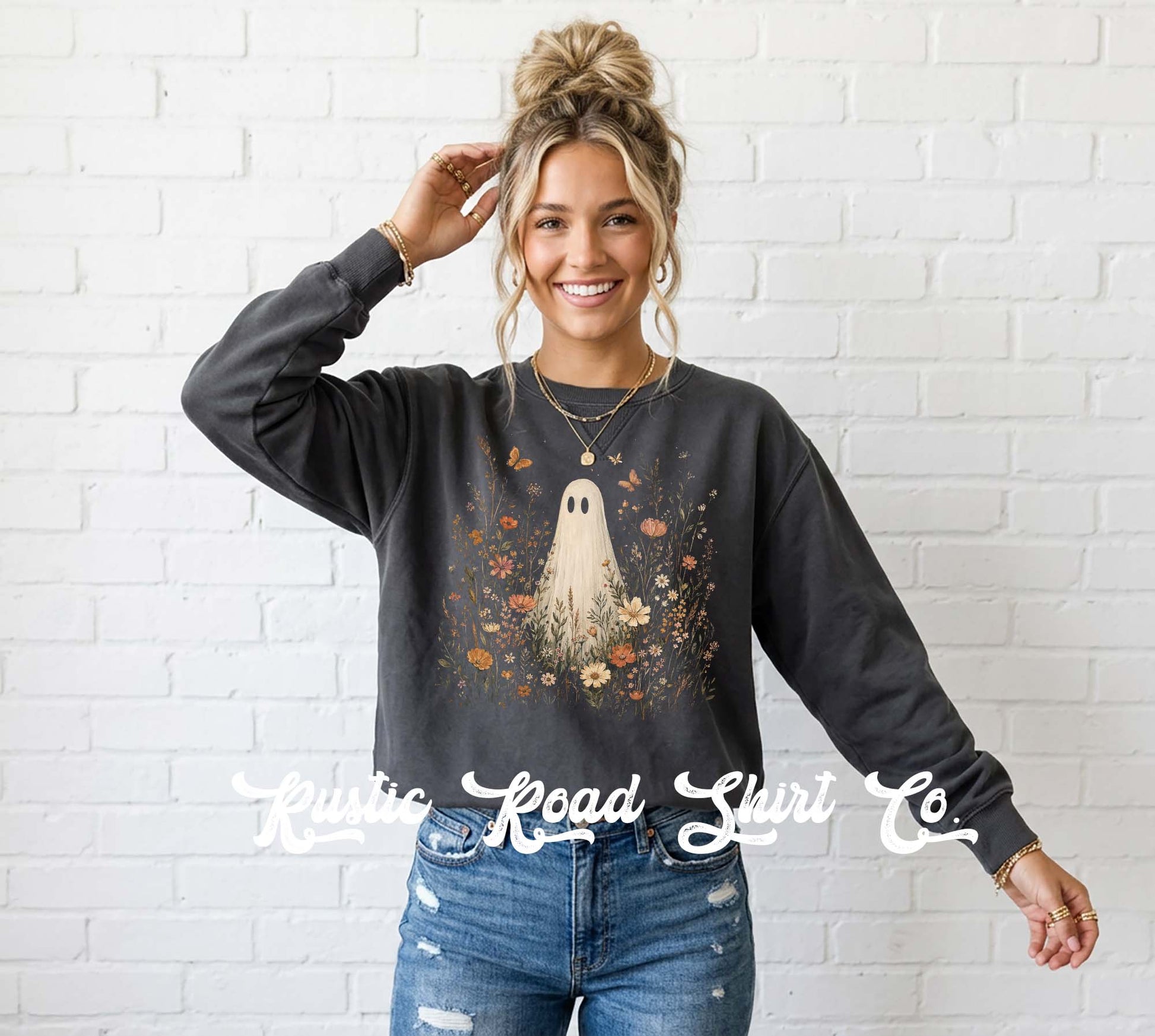 A woman wearing a dark charcoal gray crewneck sweatshirt with a ghost and floral graphic, paired with blue jeans, standing against a white brick wall. A watermark for 'Rustic Road Shirt Co' is visible across the bottom.