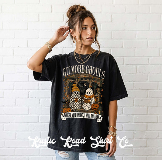 A woman stands against a white brick wall wearing a black graphic T-shirt with the words 'GILMORE Ghouls' and a Halloween-themed print featuring pumpkins, ghosts, and a coven. She is also wearing light blue jeans and layered gold necklaces and bracelets.