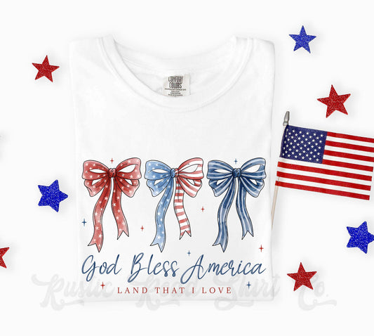 White t-shirt with red, blue, and white bows and an American flag, featuring 'God Bless America' text.