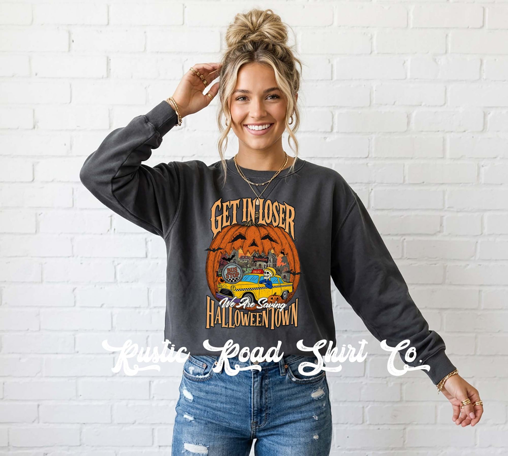 A woman wearing a dark charcoal gray sweatshirt with a Halloween-themed graphic that reads 'GET IN LOSER' and 'Halloween Town' around an orange pumpkin illustration, paired with blue jeans, standing against a white brick wall.