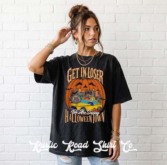A woman wearing a black graphic t-shirt with a Halloween-themed design that reads 'GET IN LOSER All We Save HALLOWEEN TOWN' around an orange jack-o'-lantern and yellow car illustration, standing against a white brick wall.