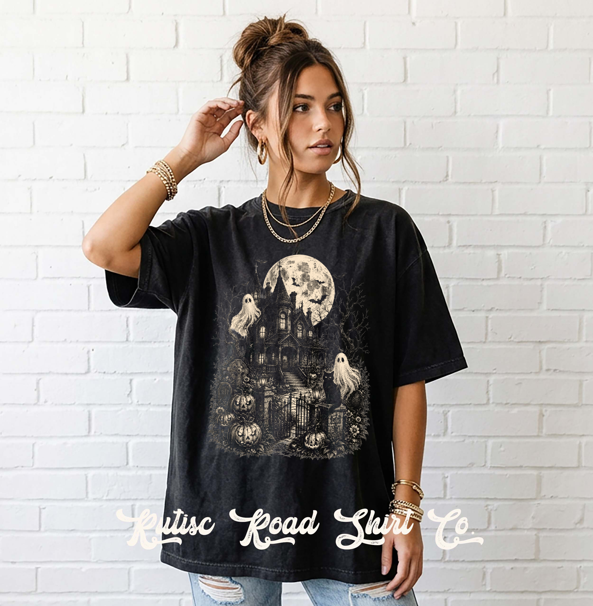 A woman wearing a black graphic t-shirt with a vintage-style Halloween print featuring a haunted house, pumpkins, ghosts, and a full moon, posed against a white brick wall. The brand name 'Kitsuc Road Shrt Co.