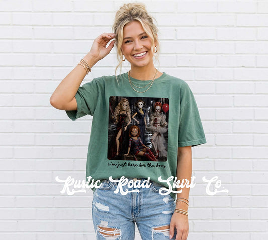 A woman wearing a washed green graphic t-shirt with a horror-themed print and the text 'I'm just here for the boys', paired with distressed blue jeans, standing against a white brick wall. A watermark for 'Rustic Road Shirts Co' is visible at the bottom.