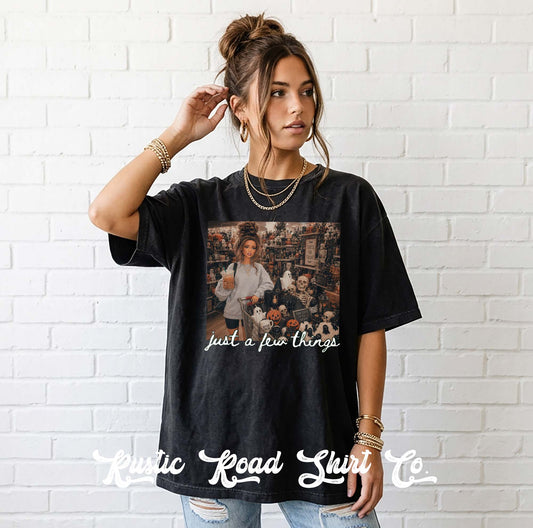 A woman models a black graphic t-shirt featuring a photo print of a band performing live music with the text 'just a few things' written below. She is standing against a white brick wall background, wearing gold jewelry and blue jeans.