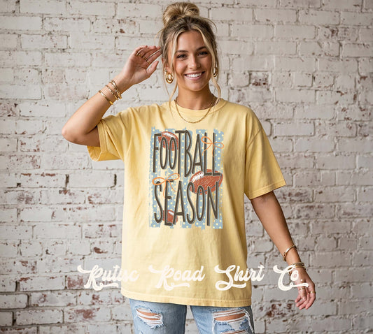 A woman wearing a pale yellow graphic t-shirt with 'FOOTBALL SEASON' printed on the front, paired with distressed blue jeans, standing against a white brick wall.