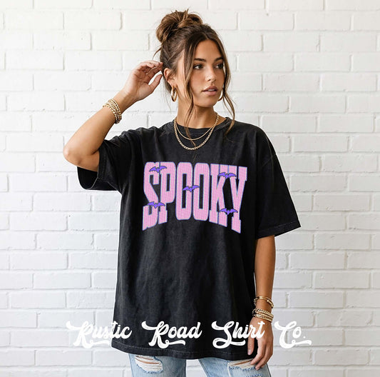 A woman wearing a black graphic t-shirt with 'SPOOKY' printed in pink and purple spiderwebs, standing against a white brick wall. The watermark 'Rustic Road Shirt Co' is visible at the bottom.