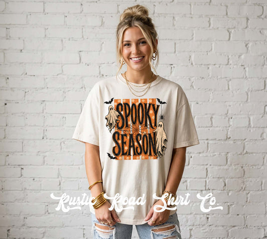 A woman wearing a cream-colored graphic t-shirt with a 'Spooky Season' Halloween design, standing against a white brick wall. The shirt features a graphic with a ghost, a pumpkin, bats, and the text 'Spooky Season' in orange and black.