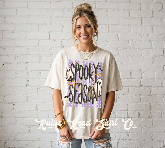 A woman wearing a cream-colored graphic t-shirt with 'Spooky Season' written in black letters over a purple patterned background, paired with distressed blue jeans, standing against a white brick wall.