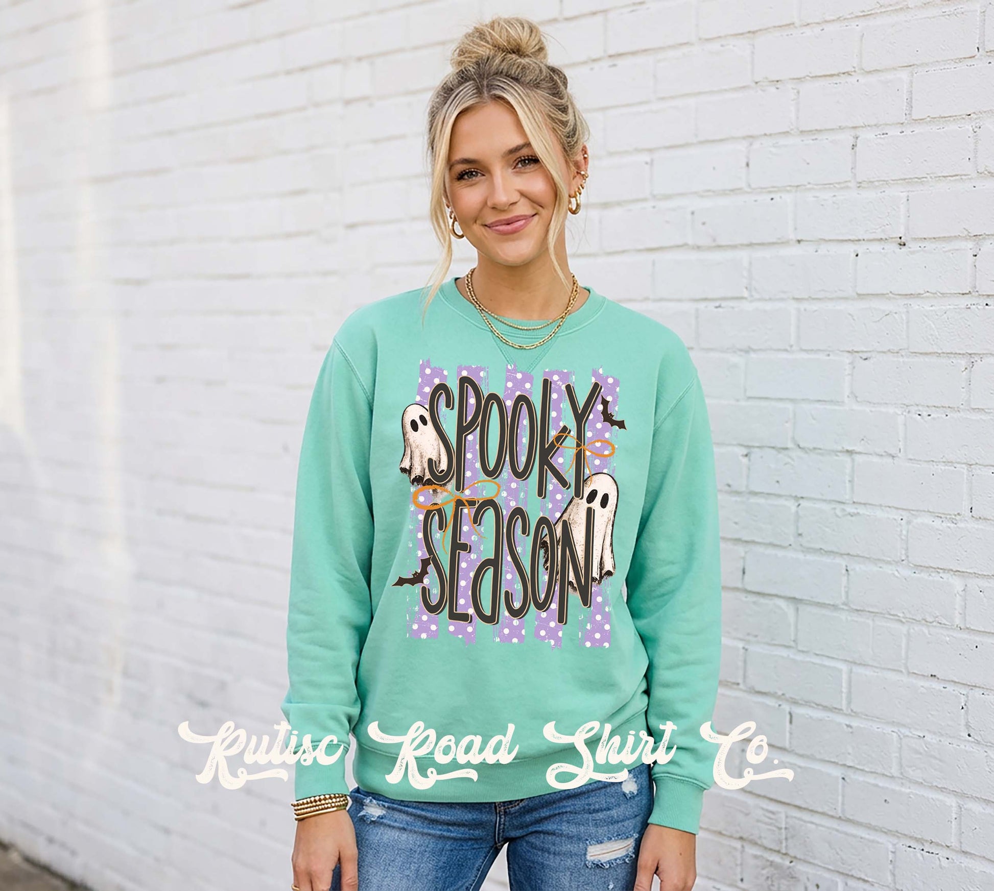 A woman wearing a mint green crewneck sweatshirt with a 'Spooky Season' Halloween graphic, standing against a white brick wall. The graphic features purple polka dots, ghosts, and bats.