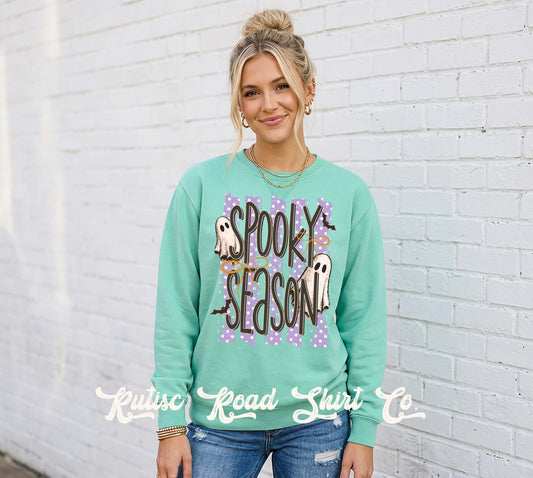 A woman wearing a mint green crewneck sweatshirt with a 'Spooky Season' Halloween graphic, standing against a white brick wall. The graphic features purple polka dots, ghosts, and bats.