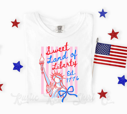 White t-shirt with 'Sweet Land of Liberty' design featuring the Statue of Liberty, American flag, and stars on a white background.