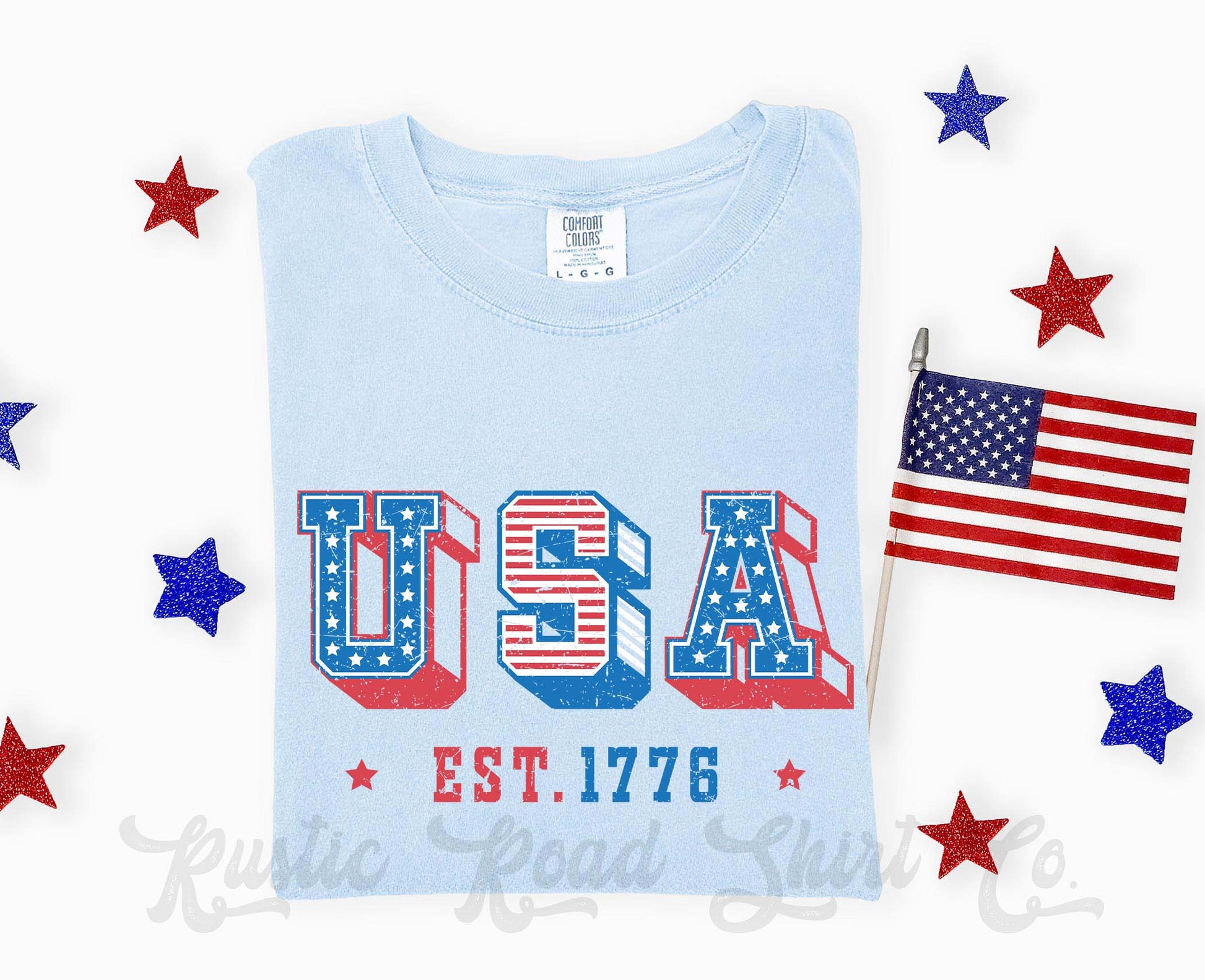 Light blue t-shirt with 'USA EST. 1776' design and American flag, surrounded by decorative stars and a small American flag.