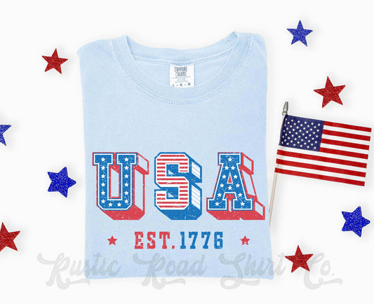 Light blue t-shirt with 'USA EST. 1776' design and American flag, surrounded by decorative stars and a small American flag.