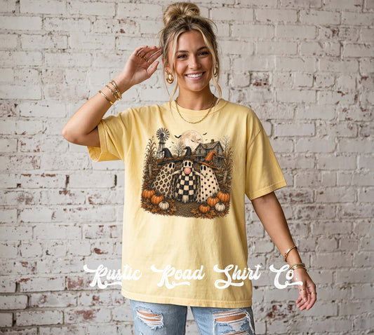 A woman wearing a pale yellow graphic t-shirt with a spooky pumpkin patch design, standing against a white brick wall. The t-shirt features a graphic of a witch's house, pumpkins, and a black-and-white checkered pumpkin.