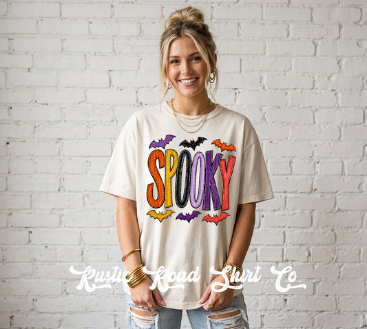 A woman wearing a cream-colored graphic t-shirt with 'SPOOKY' written in colorful letters and bats, standing against a white brick wall. A watermark for 'Krusty Road Shirt Co' is visible at the bottom.